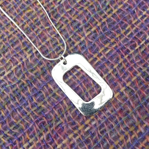 Op de afbeelding: Een zilveren ketting met een rechthoekige hanger met een uitsparing in de vorm van een rechthoek. De hanger hangt aan een zilveren ketting.