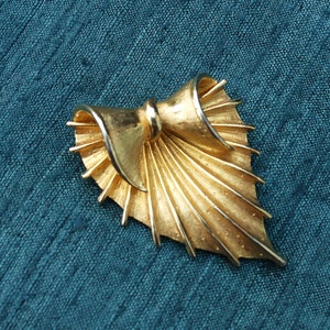 May include: A gold-toned brooch in the shape of a stylized bow with a fan-like design. The brooch is made of metal and has a textured surface.