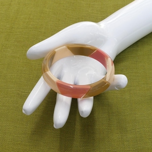 Könnte beinhalten: Ein mehrfarbiger Armreif mit rosa, beige und braunen Tönen. Der Armreif ist aus Kunststoff und hat eine glatte, glänzende Oberfläche.