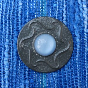 May include: A gray metal button with a star-shaped design and a light blue glass center. The button is on a blue and white woven fabric.