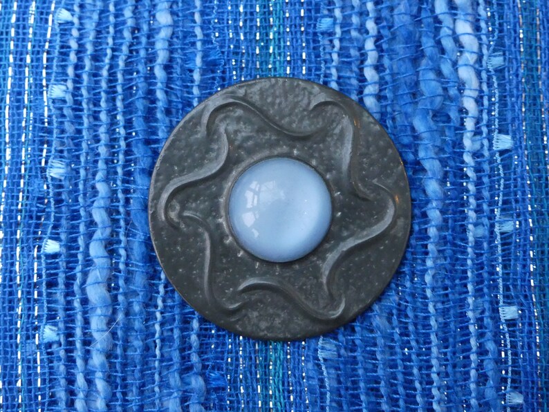 May include: A gray metal button with a star-shaped design and a light blue glass center. The button is on a blue and white woven fabric.