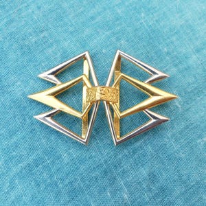 Pode incluir: Um fecho de metal decorativo com um design geométrico. O fecho apresenta elementos dourados e prateados dispostos simetricamente. A parte central tem um design em relevo, sobre um fundo de tecido azul.