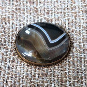 May include: A round, brown agate gemstone with white and black stripes is set in a brass bezel. The stone is surrounded by a decorative, rope-like border.