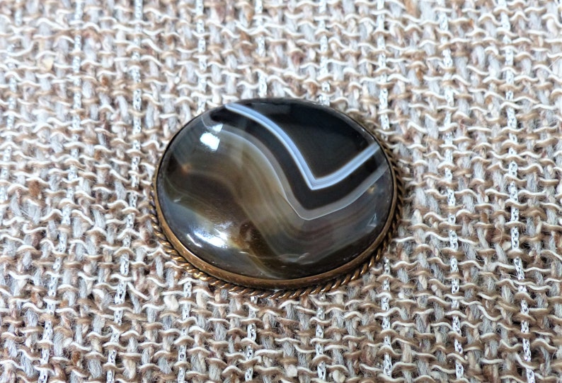 May include: A round, brown agate gemstone with white and black stripes is set in a brass bezel. The stone is surrounded by a decorative, rope-like border.