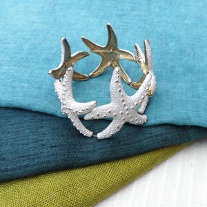 Può includere: Un braccialetto d'argento con un design di diverse stelle marine bianche. Il braccialetto è su una pila di tessuto in tonalità di blu e verde.