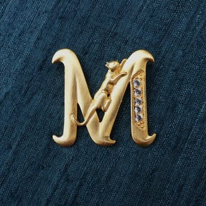 Może przedstawiać: Złota metalowa litera "M" z małym kotem wspinającym się po niej. Prawa strona litery jest ozdobiona przezroczystymi kryształami. Tło to teksturowana, ciemnoturkusowa tkanina.