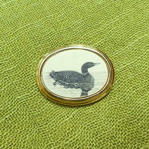 May include: An oval-shaped brooch featuring a gold-toned metal frame surrounding a central white inlay. The inlay displays a detailed black line drawing of a loon swimming on water, capturing the bird's distinct plumage and profile in a classic style.