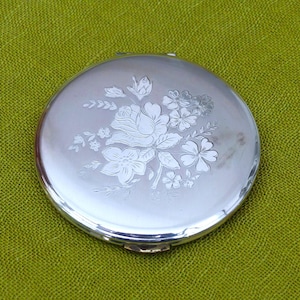 Puede incluir: Un espejo compacto redondo y plateado con un diseño floral grabado. El diseño presenta una rosa y otras flores. El compacto está sobre una superficie verde texturizada.