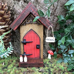 Könnte beinhalten: Ein Miniatur-Feenhaus aus Holz mit einer roten Tür, einem Willkommensschild und zwei kleinen Milchflaschen. Das Haus steht vor einem Hintergrund aus grünem Moos und einem Baumstamm.