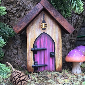 May include: A miniature wooden fairy house with a pink door, black hinges, and a yellow lantern hanging above the door. The house is sitting on a bed of moss and pine needles.