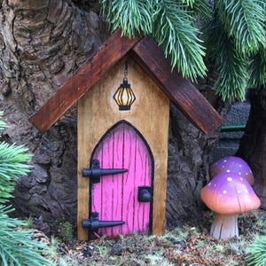 Könnte beinhalten: Ein Miniatur-Feenhaus aus Holz mit einer rosafarbenen Tür, schwarzen Scharnieren und einer kleinen schwarzen Laterne über der Tür. Das Haus steht in einem Garten mit grünem Laub und einem violetten Pilz.