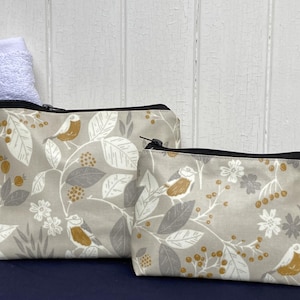 Könnte beinhalten: Zwei Reißverschlusstaschen mit einem von der Natur inspirierten Muster. Die Taschen haben einen hellgrauen Hintergrund mit einem sich wiederholenden Design von Vögeln, Blättern und Beeren in Weiß-, Grau- und Goldtönen. Eine Tasche ist größer als die andere.