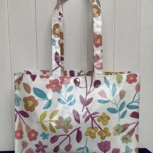 Könnte beinhalten: Weiße Tragetasche mit einem farbenfrohen Blumenmuster mit rosa, gelben, blauen und lila Blumen und grünen Blättern. Die Tasche hat passende Griffe und einen Druckknopfverschluss. Eine stilvolle und wiederverwendbare Einkaufstasche.