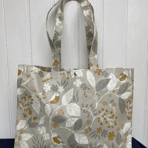 May include: A grey tote bag with a botanical print featuring birds, leaves, and berries in white, grey, and gold. The bag has two handles and a snap closure. The tote bag is made of fabric.