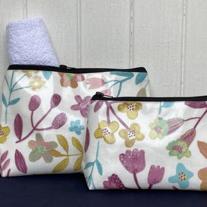 May include: Two white zippered pouches with a floral pattern in shades of pink, yellow, and blue. The larger pouch is slightly open and has a white towel inside.