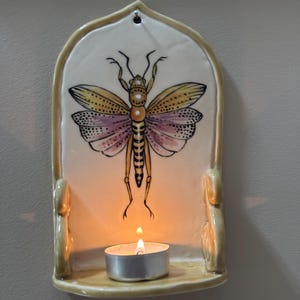 May include: A cream-colored ceramic wall sconce featuring a hand-painted illustration of a grasshopper with yellow and purple wings. The arched piece includes a small shelf at the base holding a lit tea light candle in a silver metal cup.