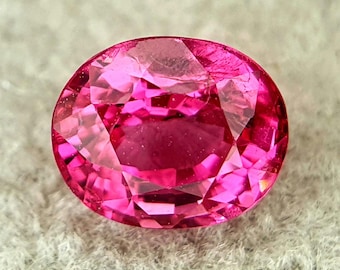 May include: A faceted, oval-shaped, hot pink gemstone. The gem has a brilliant cut with multiple facets that reflect light, creating a sparkling effect. The colour is a vibrant, saturated pink.