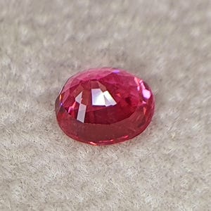 May include: A vibrant, oval-cut gemstone with a deep pinkish-red hue. The stone features a faceted surface that reflects light, creating bright highlights and internal depth across its polished, translucent, and rounded crystalline structure.