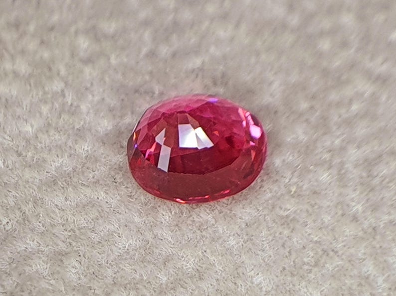 May include: A vibrant, oval-cut gemstone with a deep pinkish-red hue. The stone features a faceted surface that reflects light, creating bright highlights and internal depth across its polished, translucent, and rounded crystalline structure.