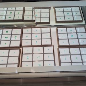 May include: A glass display case containing multiple brown wooden trays, each divided into square compartments. Inside these compartments are small, loose green gemstones, primarily emeralds, resting on white square display cards.