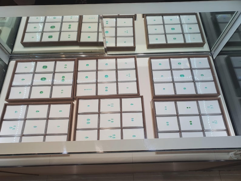 May include: A glass display case containing multiple brown wooden trays, each divided into square compartments. Inside these compartments are small, loose green gemstones, primarily emeralds, resting on white square display cards.