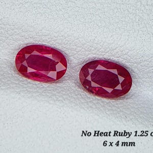 Op de afbeelding: Twee ovale, rode robijnen, elk 6 x 4 mm groot. De robijnen liggen op een wit oppervlak. De tekst "No Heat Ruby 1.25 ct 6 x 4 mm" staat onder de robijnen.