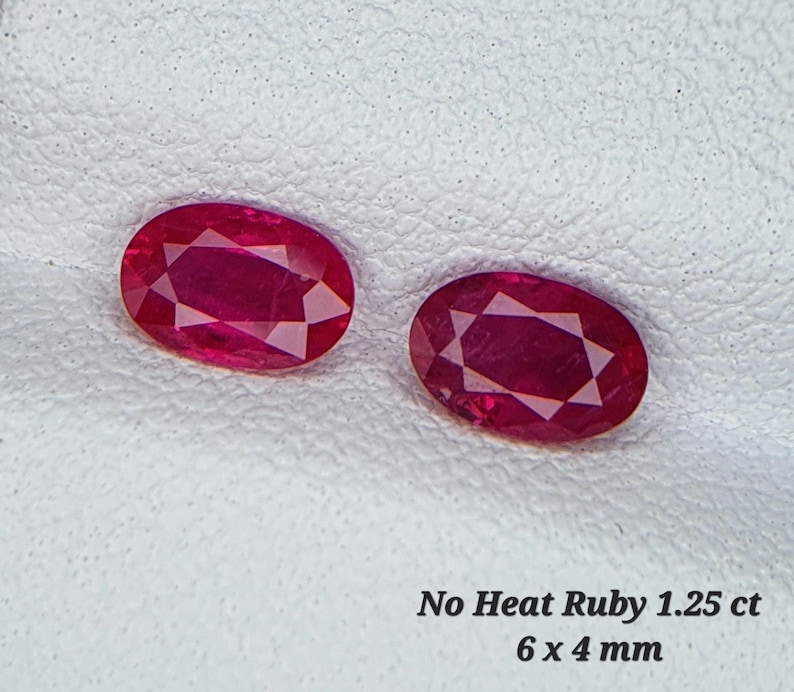 Op de afbeelding: Twee ovale, rode robijnen, elk 6 x 4 mm groot. De robijnen liggen op een wit oppervlak. De tekst "No Heat Ruby 1.25 ct 6 x 4 mm" staat onder de robijnen.