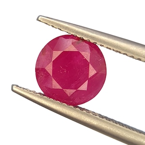 May include: A faceted, round ruby gemstone held by silver tweezers. The ruby is a vibrant, deep red color with a star-like pattern visible in the center. The gemstone is set against a clean, white background.