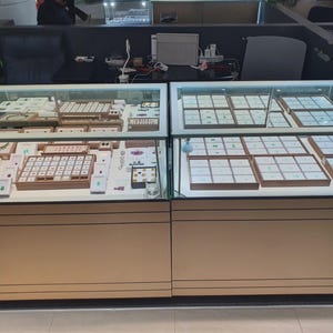 May include: Two side-by-side glass jewellery display cases with light wood-toned bases. The cases contain multiple white trays organised into grids, holding various loose gemstones and finished jewellery pieces under bright interior lighting.