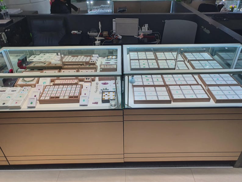 May include: Two side-by-side glass jewellery display cases with light wood-toned bases. The cases contain multiple white trays organised into grids, holding various loose gemstones and finished jewellery pieces under bright interior lighting.