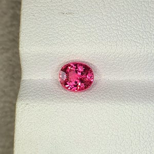 May include: A vibrant, oval-cut gemstone with a deep pink hue and multifaceted surface that reflects light. The stone features a brilliant, clear clarity and is positioned against a textured, off-white surface with a distinct horizontal fold.