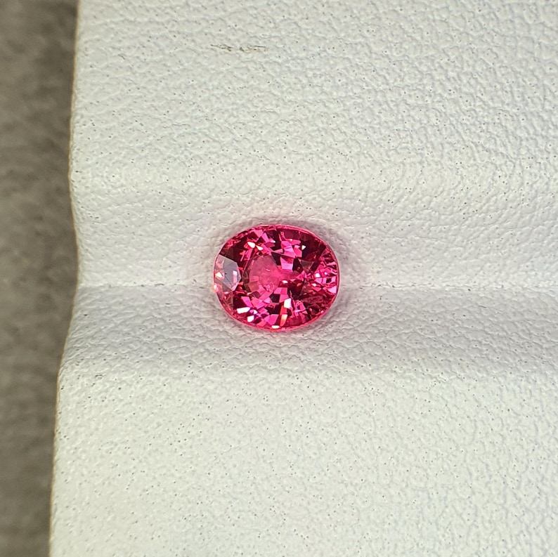 May include: A vibrant, oval-cut gemstone with a deep pink hue and multifaceted surface that reflects light. The stone features a brilliant, clear clarity and is positioned against a textured, off-white surface with a distinct horizontal fold.