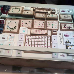 May include: A glass display case filled with an assortment of jewellery, including necklaces, rings, and loose gemstones arranged in beige and white trays. The collection features various coloured stones, including green, red, and blue, set in metal.