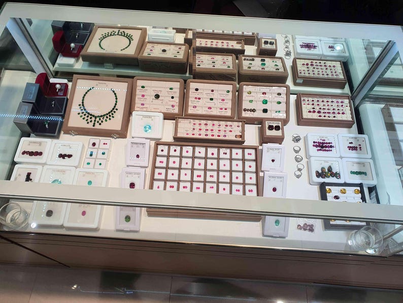 May include: A glass display case filled with an assortment of jewellery, including necklaces, rings, and loose gemstones arranged in beige and white trays. The collection features various coloured stones, including green, red, and blue, set in metal.