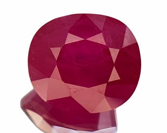 May include: A deep red, oval-cut gemstone with a faceted surface that reflects light. The stone displays a vibrant, saturated crimson hue and a symmetrical arrangement of triangular and trapezoidal facets across its top and side profile.