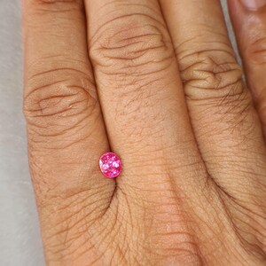 May include: A small, oval-cut gemstone with a vibrant, saturated pink hue. The stone features a faceted surface that reflects light, creating a sparkling appearance. It is positioned against a neutral background, showcasing its clear, translucent quality and precise, symmetrical shape.