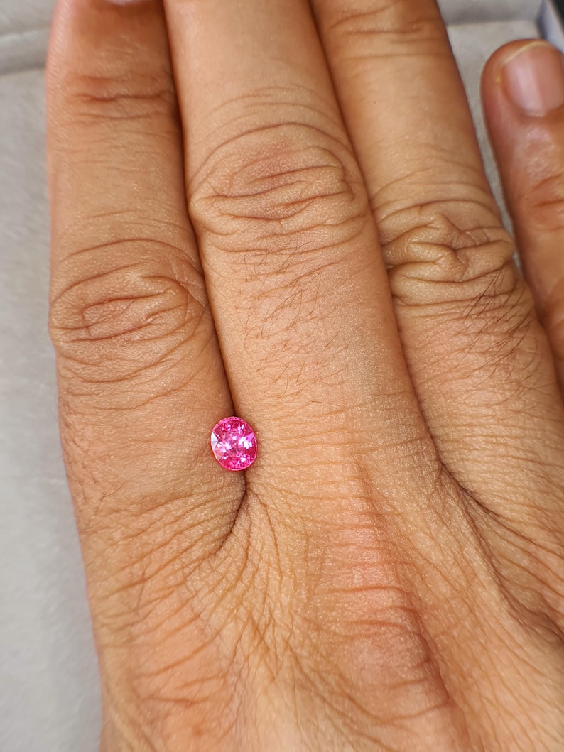 May include: A small, oval-cut gemstone with a vibrant, saturated pink hue. The stone features a faceted surface that reflects light, creating a sparkling appearance. It is positioned against a neutral background, showcasing its clear, translucent quality and precise, symmetrical shape.
