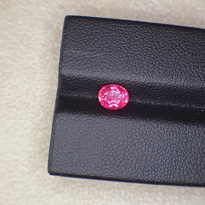 May include: A vibrant, oval-cut pink gemstone with a faceted surface that reflects light. The gemstone is positioned on a black, textured display stand that features a horizontal groove running across its centre, providing a stark contrast.