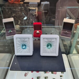 May include: Two loose Paraiba tourmaline gemstones in white display cases, each with a label. Behind them are three jewellery boxes: two grey boxes containing rings and one red box with a pearl ring. The display is inside a glass jewellery case.