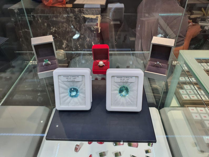 May include: Two loose Paraiba tourmaline gemstones in white display cases, each with a label. Behind them are three jewellery boxes: two grey boxes containing rings and one red box with a pearl ring. The display is inside a glass jewellery case.