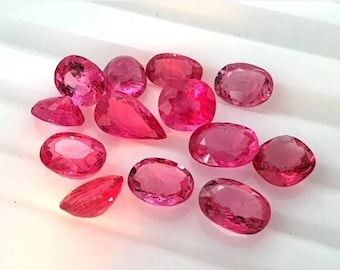 May include: A collection of twelve faceted loose gemstones in various shades of vibrant pink and deep red. The stones are cut into oval, pear, and cushion shapes, each displaying internal light reflection and clear, polished surfaces throughout.
