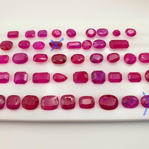 May include: A selection of deep red rubies in various shapes, presented on a white surface. The gemstones, including oval, rectangular, and round cuts, vary in size and colour intensity. Suitable for jewellery design.