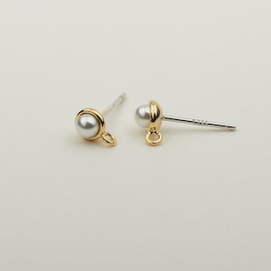 May include: A pair of gold-tone pearl stud earrings with a small loop on the bottom. The earrings are on a white background.