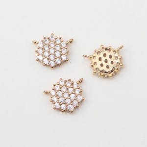May include: Three gold-colored, round jewelry charms. Each charm is encrusted with small, clear, sparkling stones. The charms have small loops on either side for attaching to other jewelry pieces. The charms are on a light-colored background.