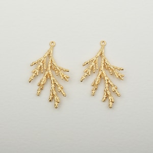 Puede incluir: Dos colgantes dorados en forma de hoja con una superficie texturizada. Cada colgante tiene un pequeño lazo en la parte superior para sujetarlo a la joyería. Los colgantes están diseñados para parecerse a una ramita de planta.