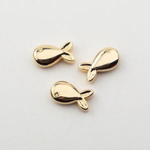 May include: Three gold-coloured fish-shaped charms. Each charm has a rounded body, a small eye detail, and a three-lobed tail. The charms are arranged on a light background, showcasing their shiny, reflective surface.