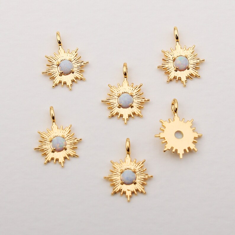 May include: Gold-colored sunburst pendants with a central, light-colored stone. The pendants have a detailed, radiant design with a loop at the top for attaching to a chain or other jewelry. The background is a plain, light color.