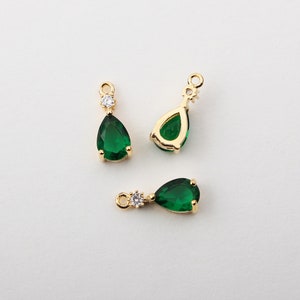 May include: Three gold-colored teardrop-shaped pendants with emerald green gemstones. Each pendant has a small, round, clear gemstone and a loop for attaching to a chain or earring hook. The pendants are on a white background.