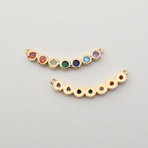以下が含まれることがあります： 2つの金色の曲線的なジュエリーチャーム。各チャームは、虹色の配置で7つのカラフルな丸い宝石の列を備えています。宝石には、赤、オレンジ、黄色、緑、青、紫が含まれます。チャームの両端には小さなループが付いています。