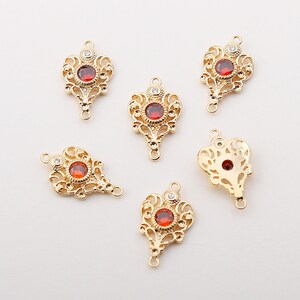 May include: Gold-coloured filigree pendants with a central red gemstone and a smaller clear stone. The pendants have an ornate design with loops for attaching to jewellery. These pendants are likely for jewellery making, with a decorative design.